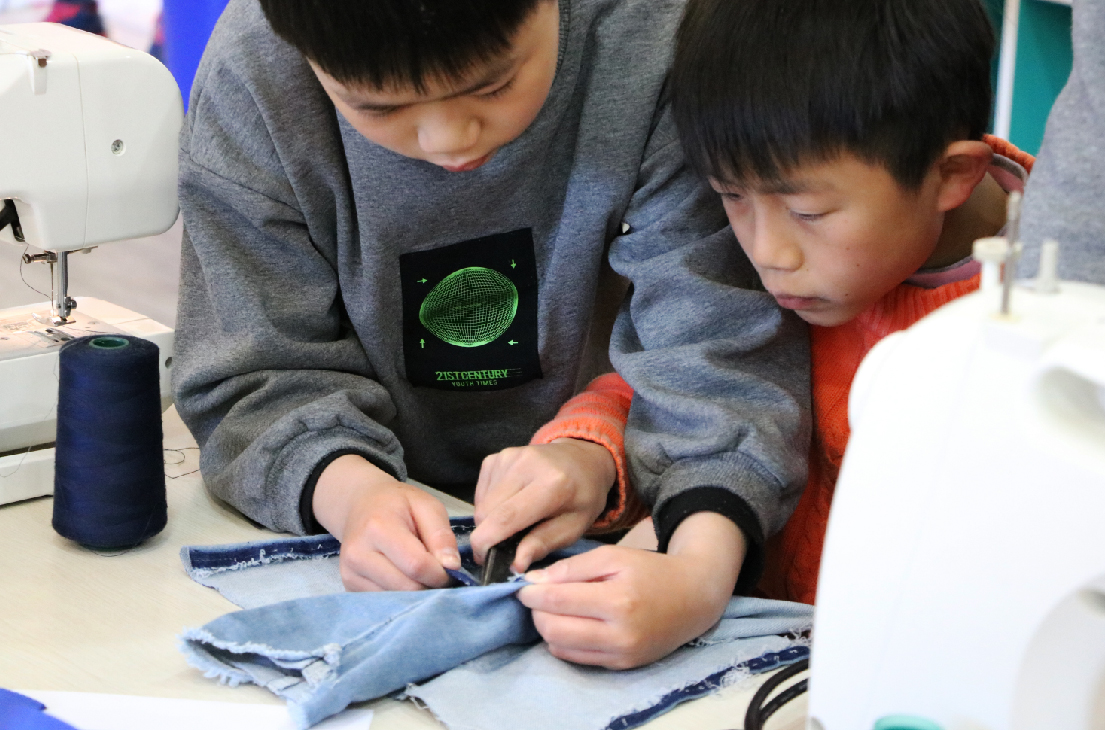 兩位年幼兒童在工作台前進行手工縫紉活動。左邊穿著灰色「21世紀技能」T恤的男童正在縫製藍色布料，右邊穿著紅色袖子的男童也參與其中。桌上放有縫紉機、深藍色線軸等工具，展示兒童進行實踐性創意學習的情景。