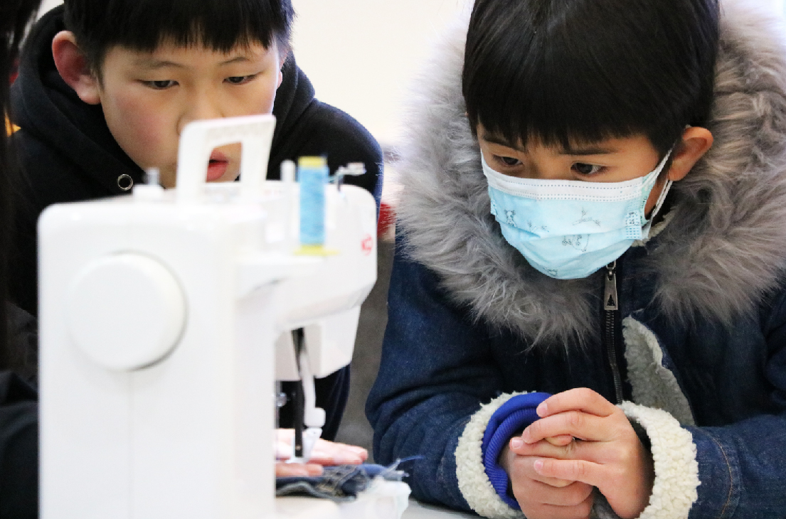 兩名佩戴口罩的兒童專注地操作一台白色迷你縫紉機，進行手工藝活動。左側男童低頭觀看機器，右側女童集中注意力操作，展現兒童學習實踐技能的教育場景。