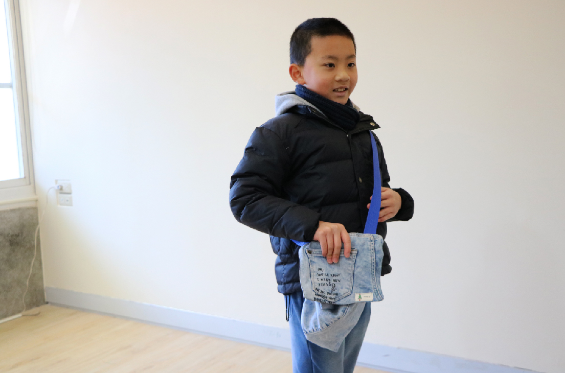 一名小男孩穿著深藍色羽絨外套，背著藍色肩帶，佩戴一個牛仔布改造的小側背包。他在明亮的室內空間轉身微笑，展示自己的手工製作包包。