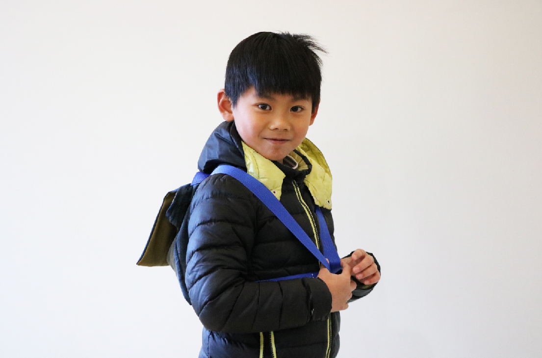 一位穿著深藍色羽絨外套、黃色領子的亞洲男童，背著藍色背帶的背包，面向鏡頭微笑，站在白色背景前的半身照。
