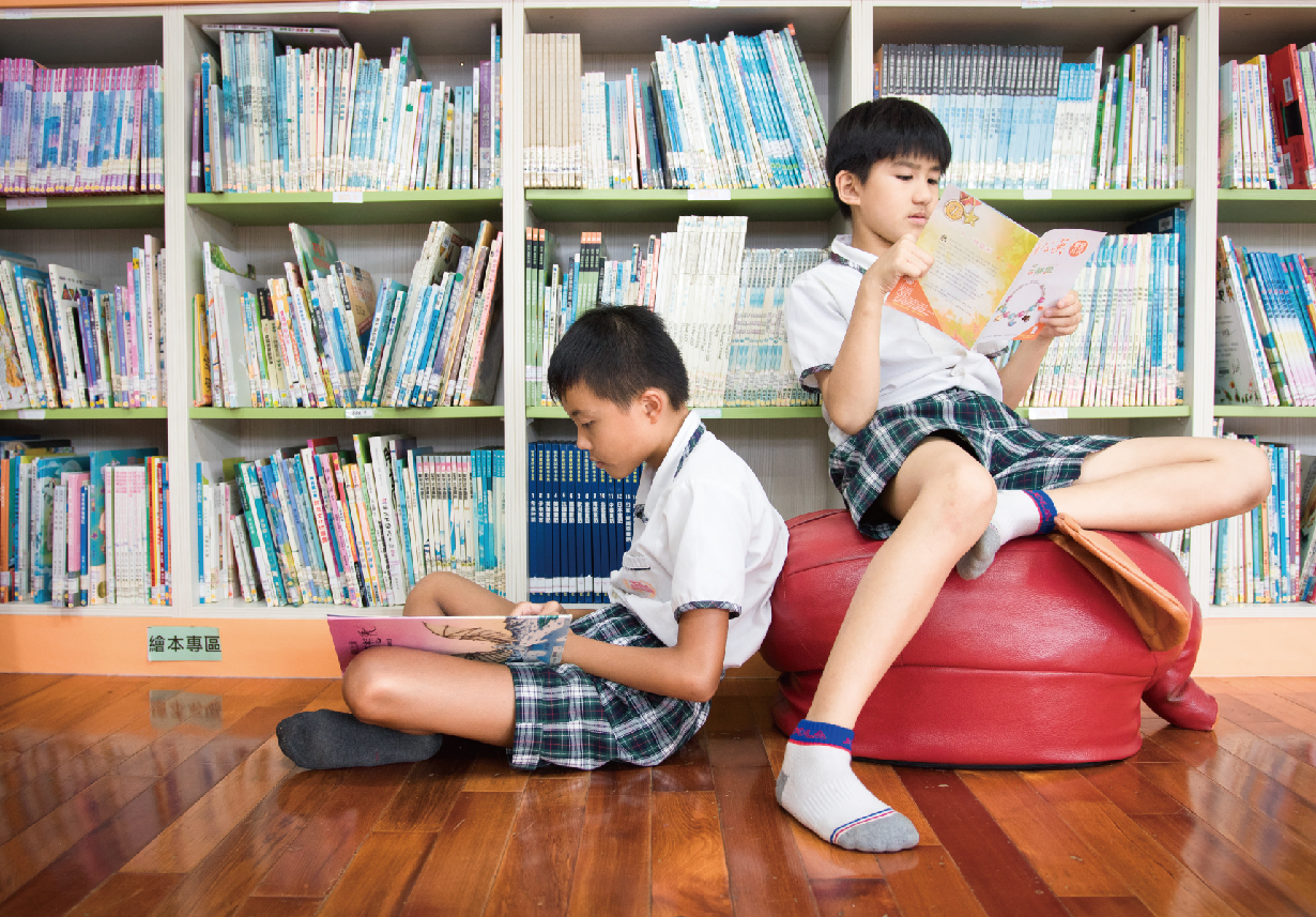 兩位穿著校服的學生在圖書館內閱讀。一人坐在地上看書，另一人坐在紅色坐墊上閱讀雜誌。身後是擺滿各類書籍的白色書架，地板為木質。標示牌寫著「繪本專區」。