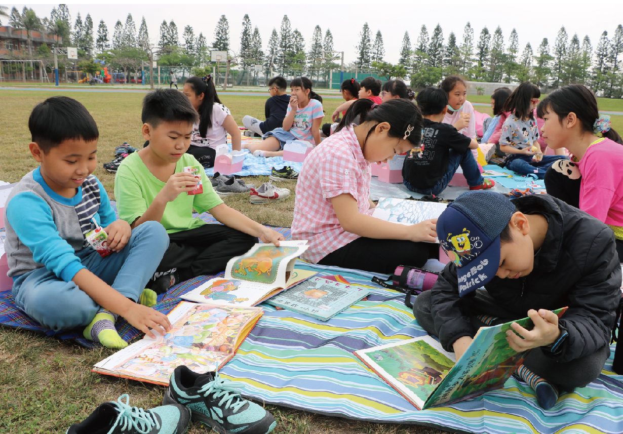 一群小學生在校園草地上圍坐成圈，坐在彩色野餐墊上閱讀兒童書籍。背景是運動場、樹木和晴朗天空。這是一場校園戶外閱讀活動，孩子們認真專注地看書。