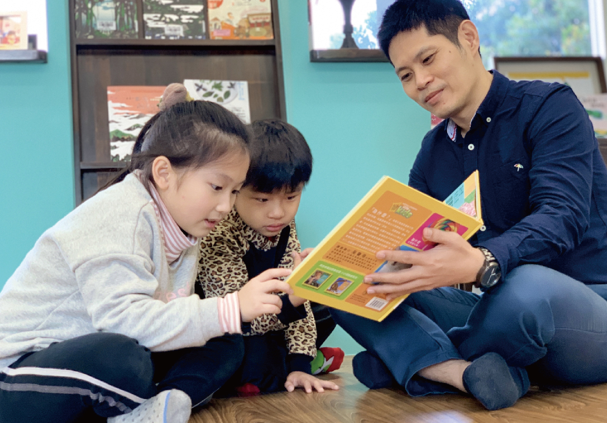 在教室內，一位女老師蹲下身與兩名小女孩一起閱讀黃色兒童圖書。背景是藍綠色牆面和展示櫃，地上鋪著木質地板。這是一個溫馨的閱讀教學場景。