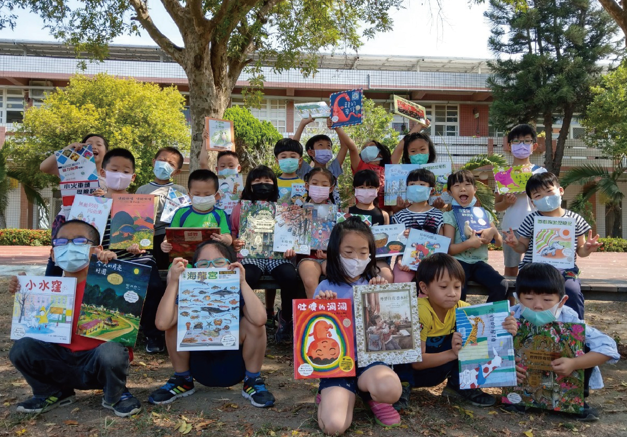 一群小學生在校園內集合拍照，他們戴著口罩，手持自己創作的繪本和書籍，站立或蹲坐在大樹下，身後是校舍建築。這是一場閱讀或創意寫作成果展示活動。