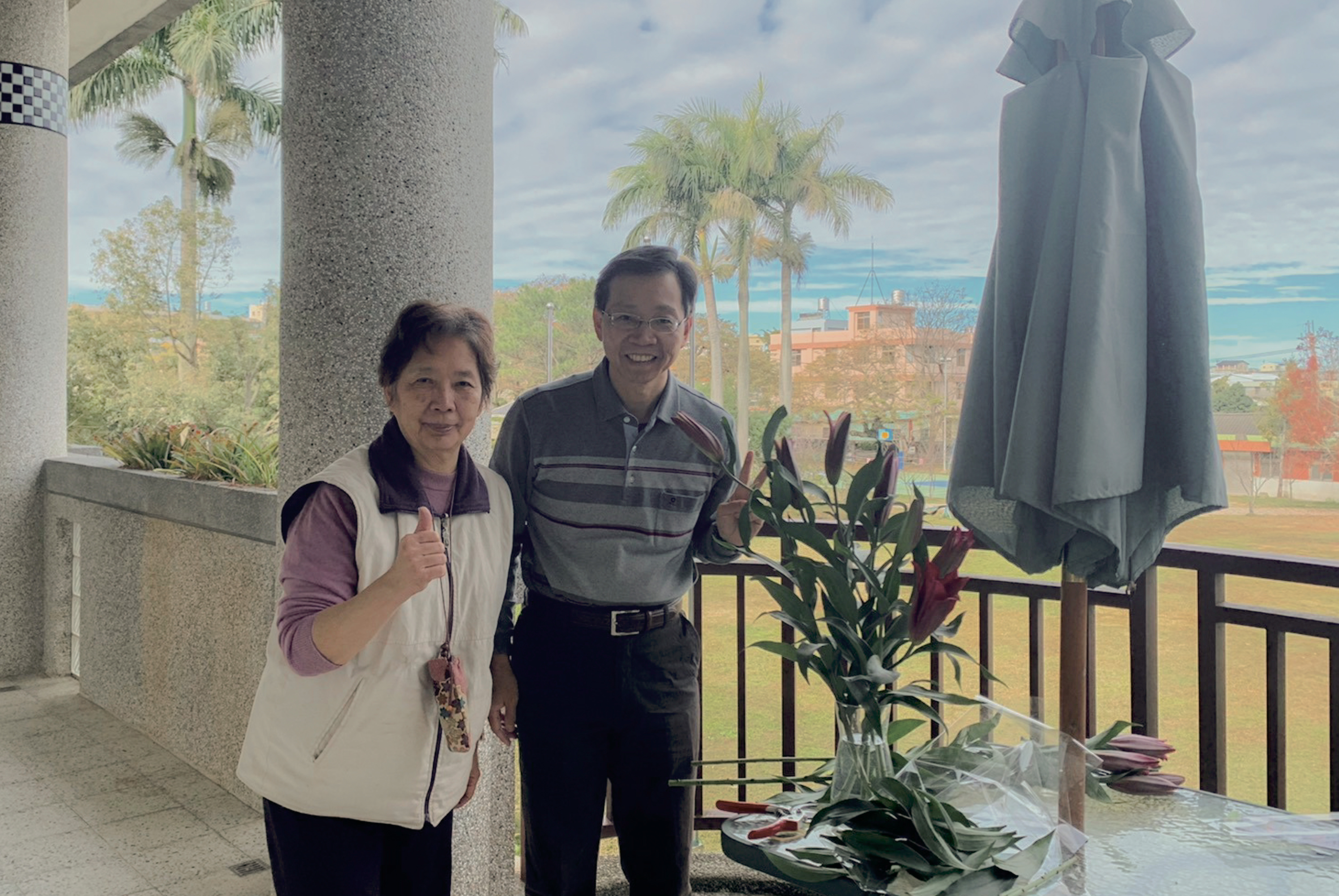 兩位亞洲遊客在地中海沿岸的露台上合影。背景是棕櫚樹、紅磚建築、藍色海景和灰色雕像。女性穿著米色背心和紫色長袖上衣，男性穿著條紋短袖襯衫。