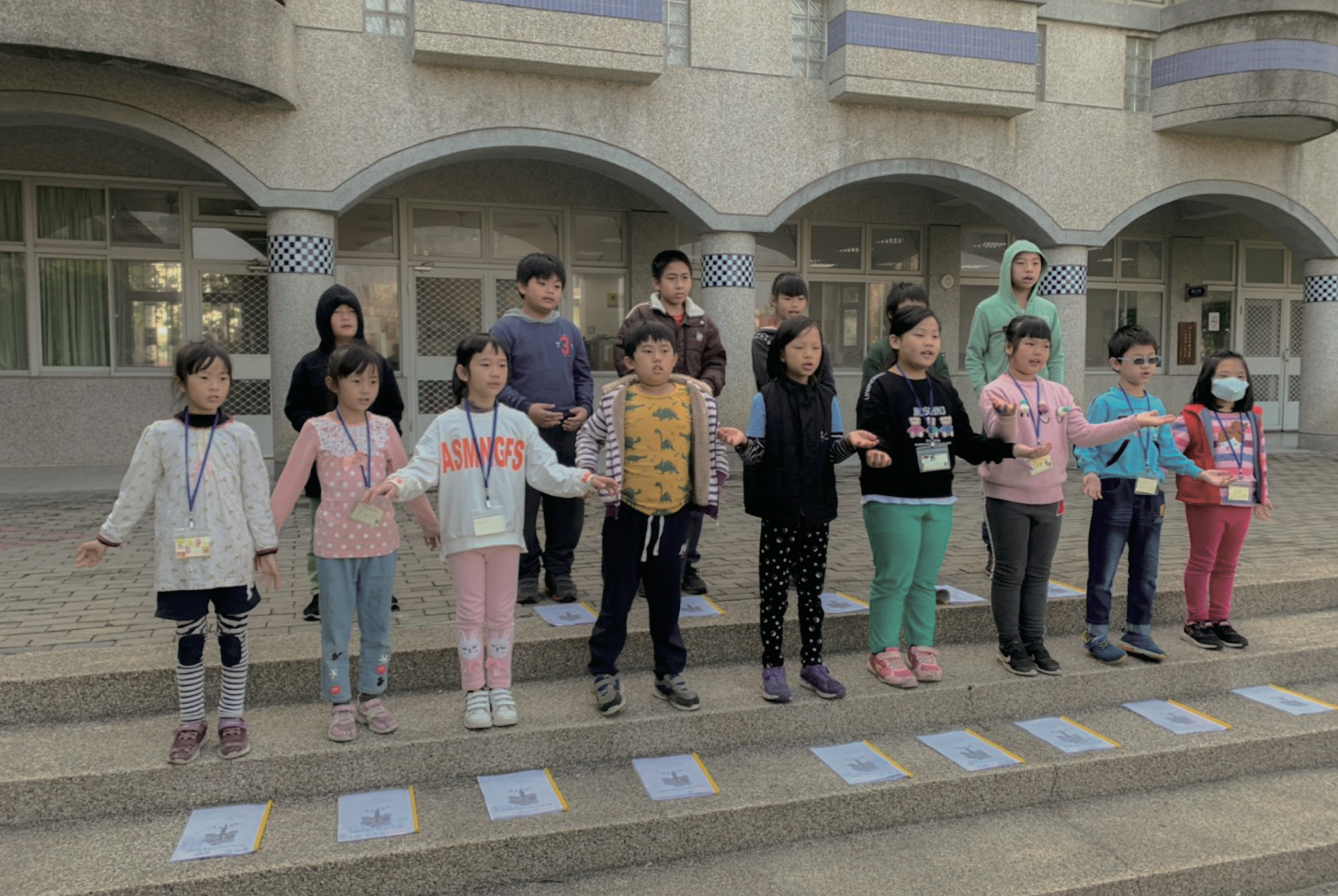 一群小學生在學校操場上排成一列，面向鏡頭。他們穿著各色冬季服裝，大多戴著識別證。地上擺放著多張藍色和黃色的卡片。背景是現代化的校舍建築，有拱形走廊和格狀欄杆。