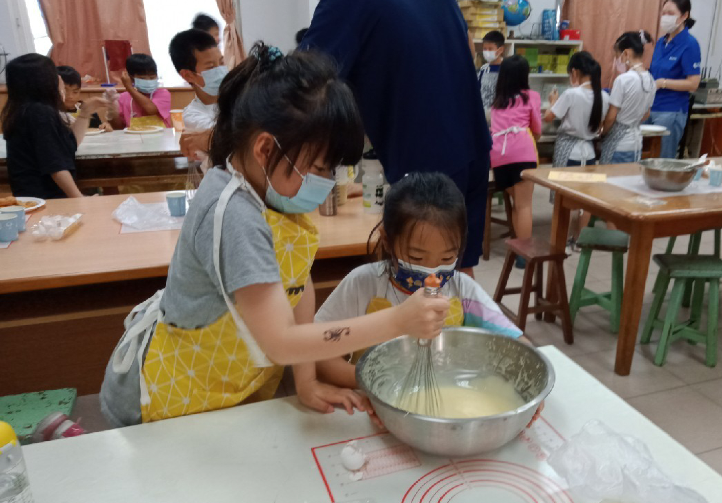 幼兒園教室內，一名戴口罩的教師指導小女孩用筷子在金屬碗中攪拌麵糊。背景有多名幼童在進行烹飪活動，牆上貼著紅色圓形圖案。這是一堂烹飪教育課程。