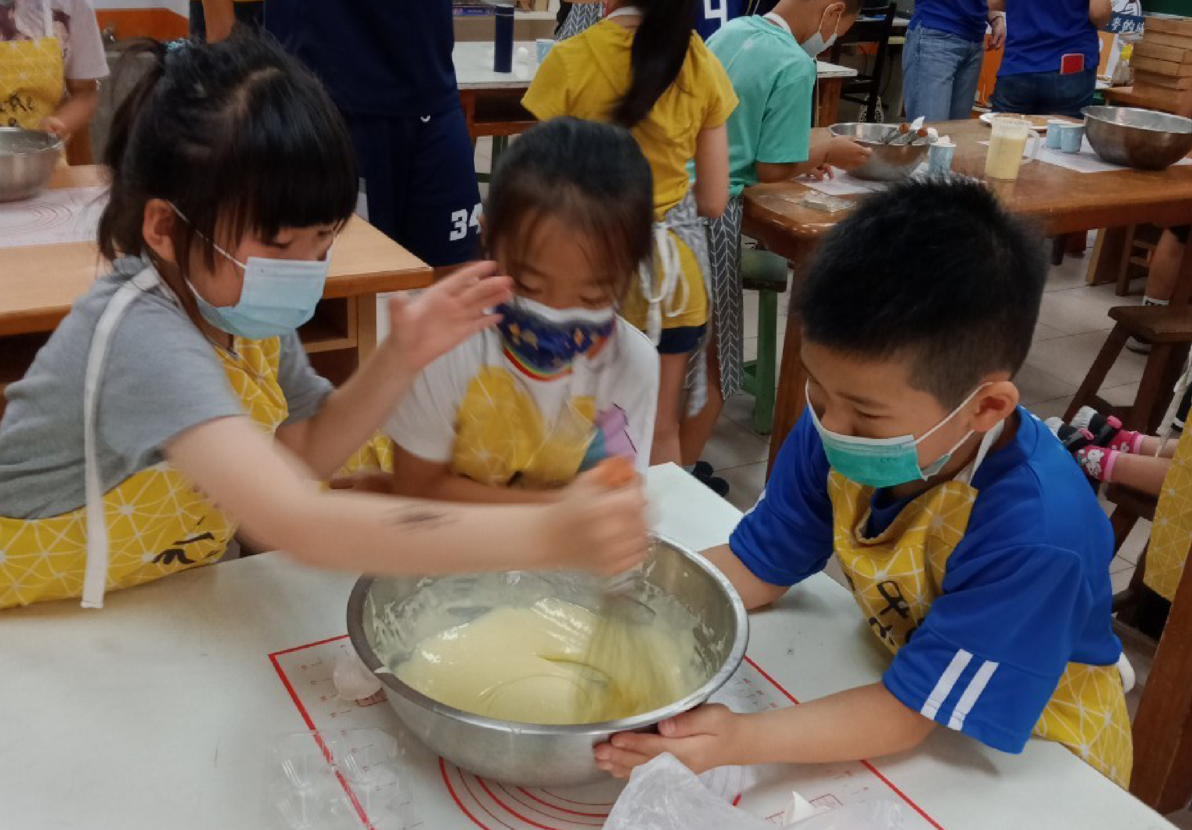 學童們穿著黃色圍裙在教室進行烹飪活動。一位女性志工協助小男孩攪拌金屬盆中的白色麵團，另一名小女孩也在旁邊參與。多名學童戴著口罩，認真參與這項動手實踐的教育活動。
