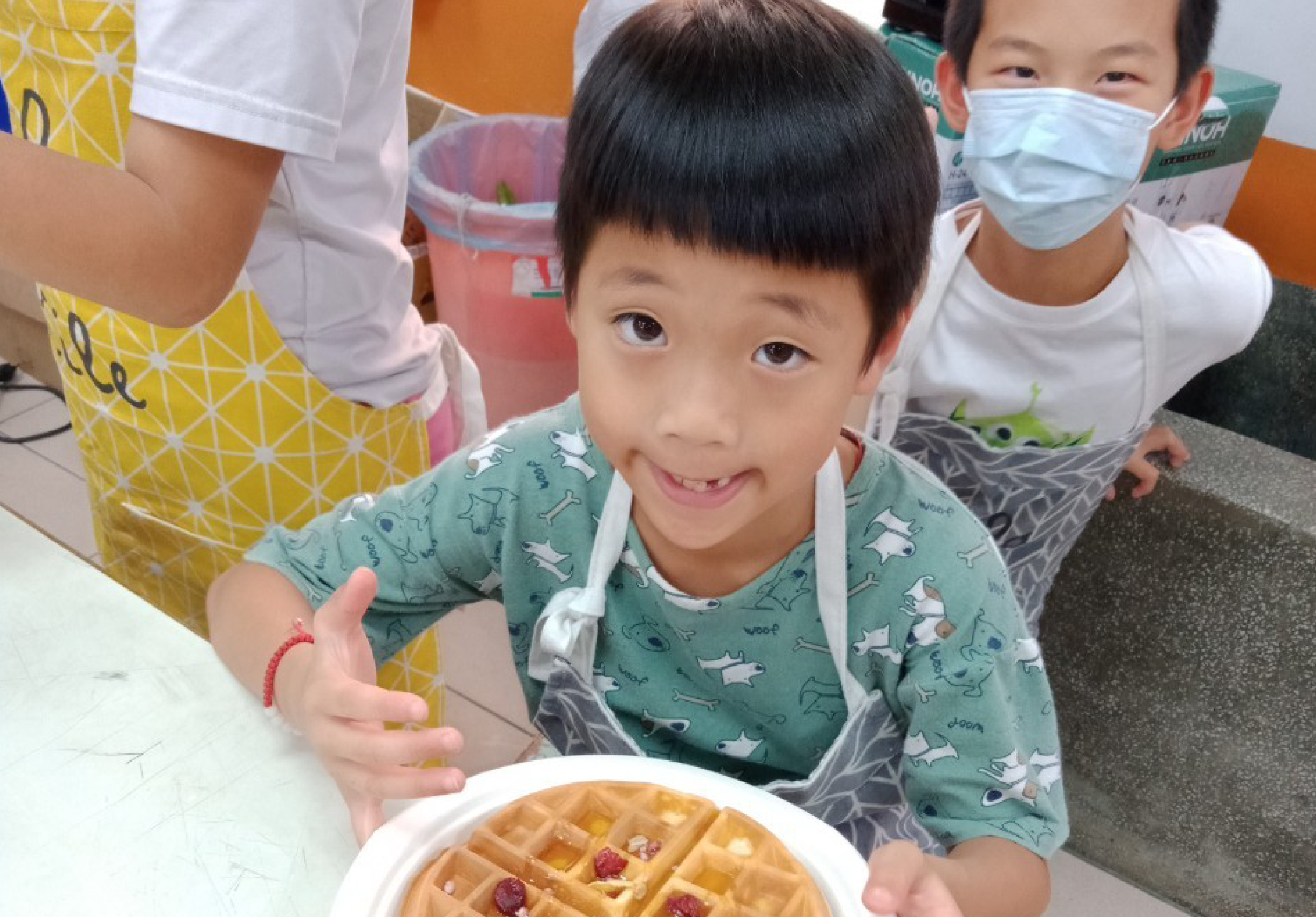 一名穿著綠色動物圖案上衣的小男孩在烹飪課上展示自己製作的鬆餅，鬆餅上裝飾著紅色莓果。身後站著穿著圍裙的成人和戴著口罩的同學，在溫暖的室內教室環境中進行烘焙活動。