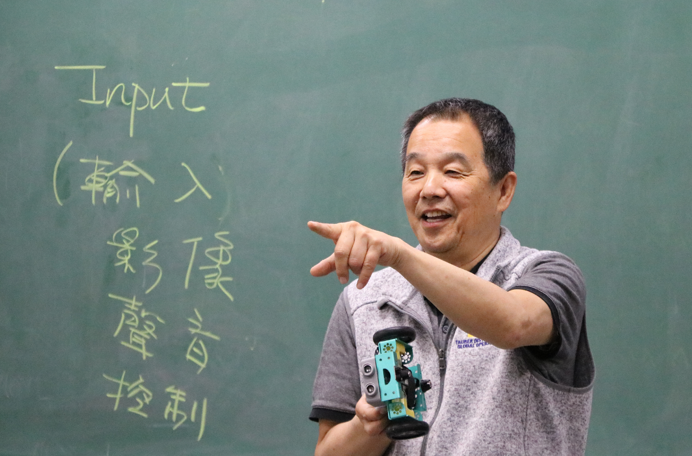 一位男性教師在綠色黑板前進行教學，黑板上以黃色粉筆寫著中文字和「Input」等教學內容。教師穿著灰色T恤，右手指向黑板，左手持藍色教學用具，表情生動，正在向學生講解課程內容。
