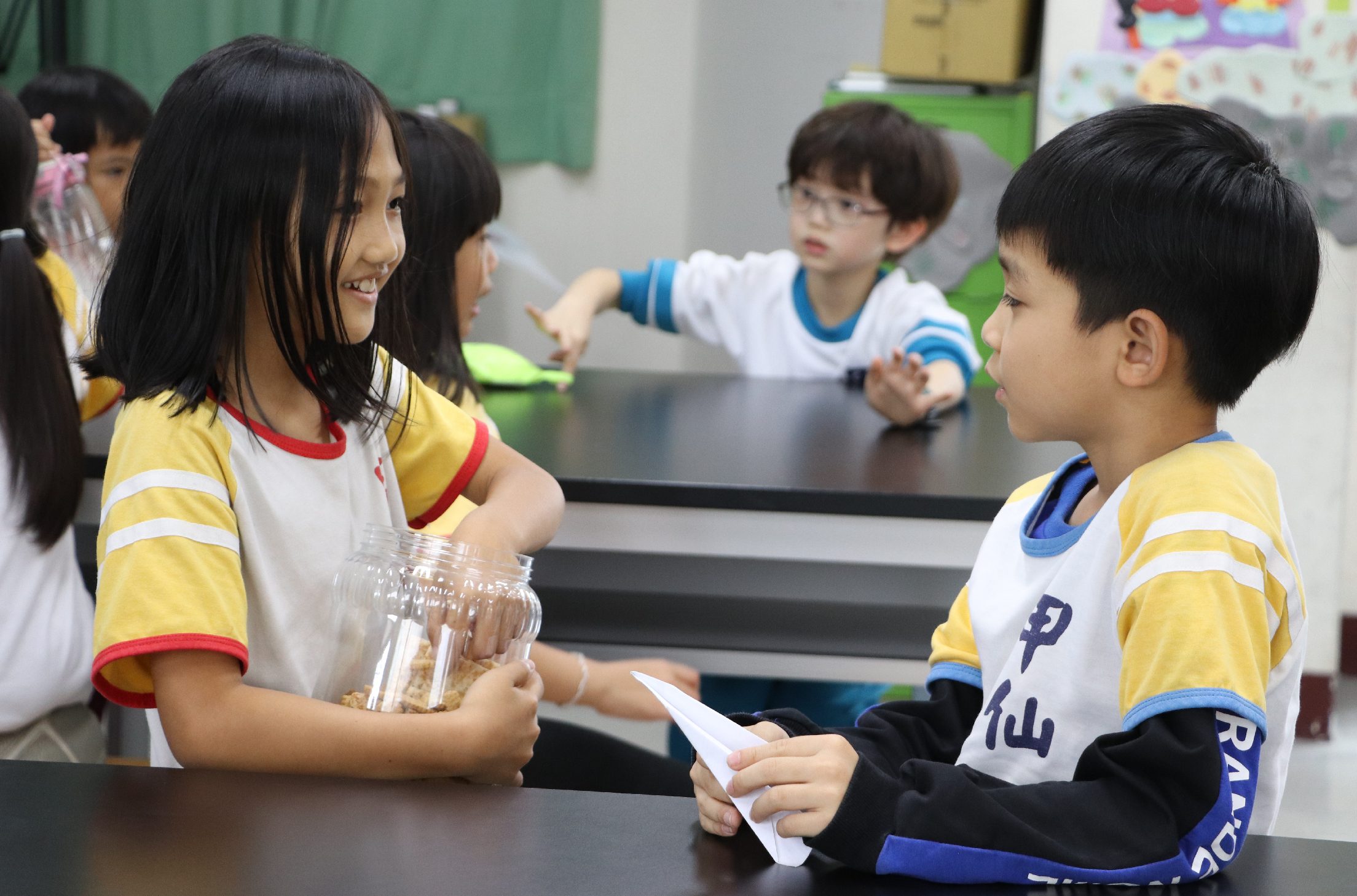 教室內，穿著黃白相間運動服的小學生們圍坐在黑色桌邊進行活動。左側女學生手持裝有穀物的玻璃罐，與右側男學生交談互動。背景可見其他學生正在進行相關活動，牆面貼有彩色海報。