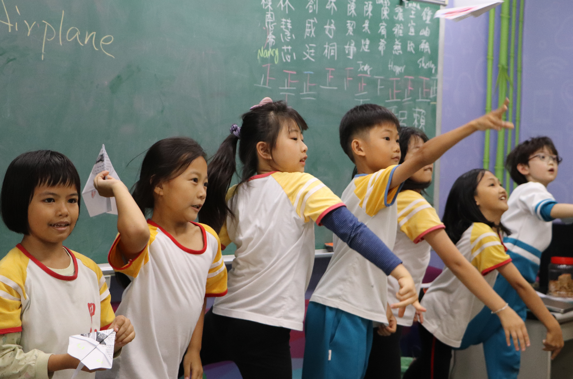 教室內，約八名穿著黃白色制服的小學生站在綠色黑板前，舉著紙飛機，做出投擲動作。他們看起來正在進行課堂活動或表演，黑板上寫滿中文字和符號。