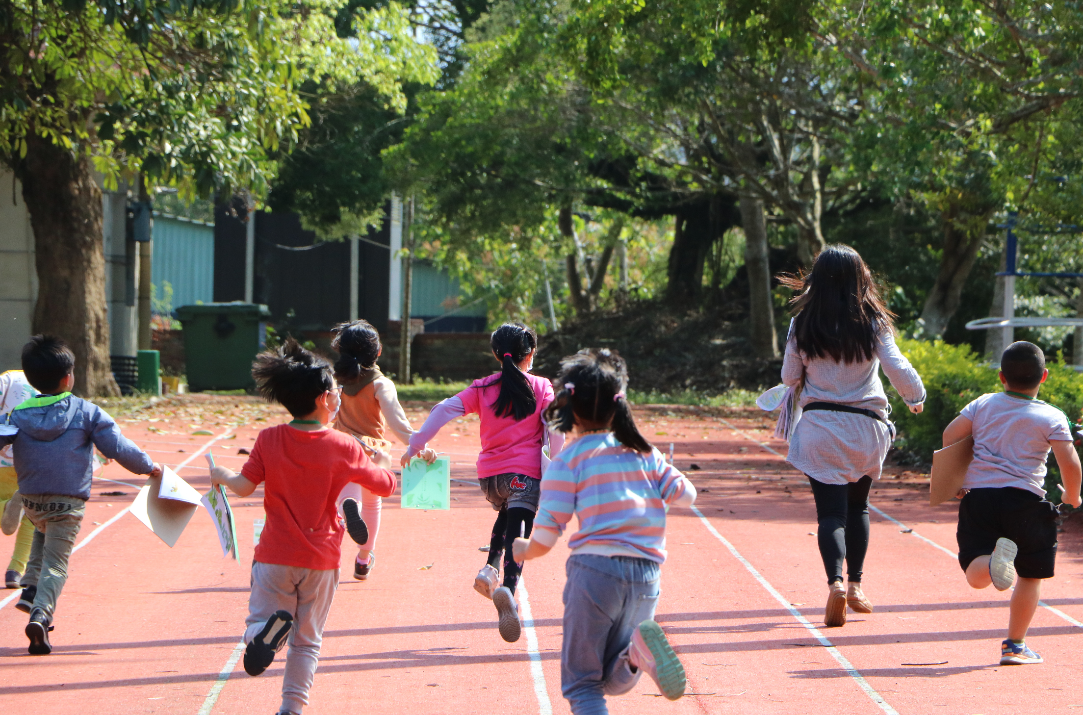 一群小學生在紅色運動場上奔跑，由一位女性教師帶隊。孩子們穿著各色衣服，在白線標記的跑道上進行體育活動。背景是茂密的樹木和學校建築，陽光灑落在場地上。