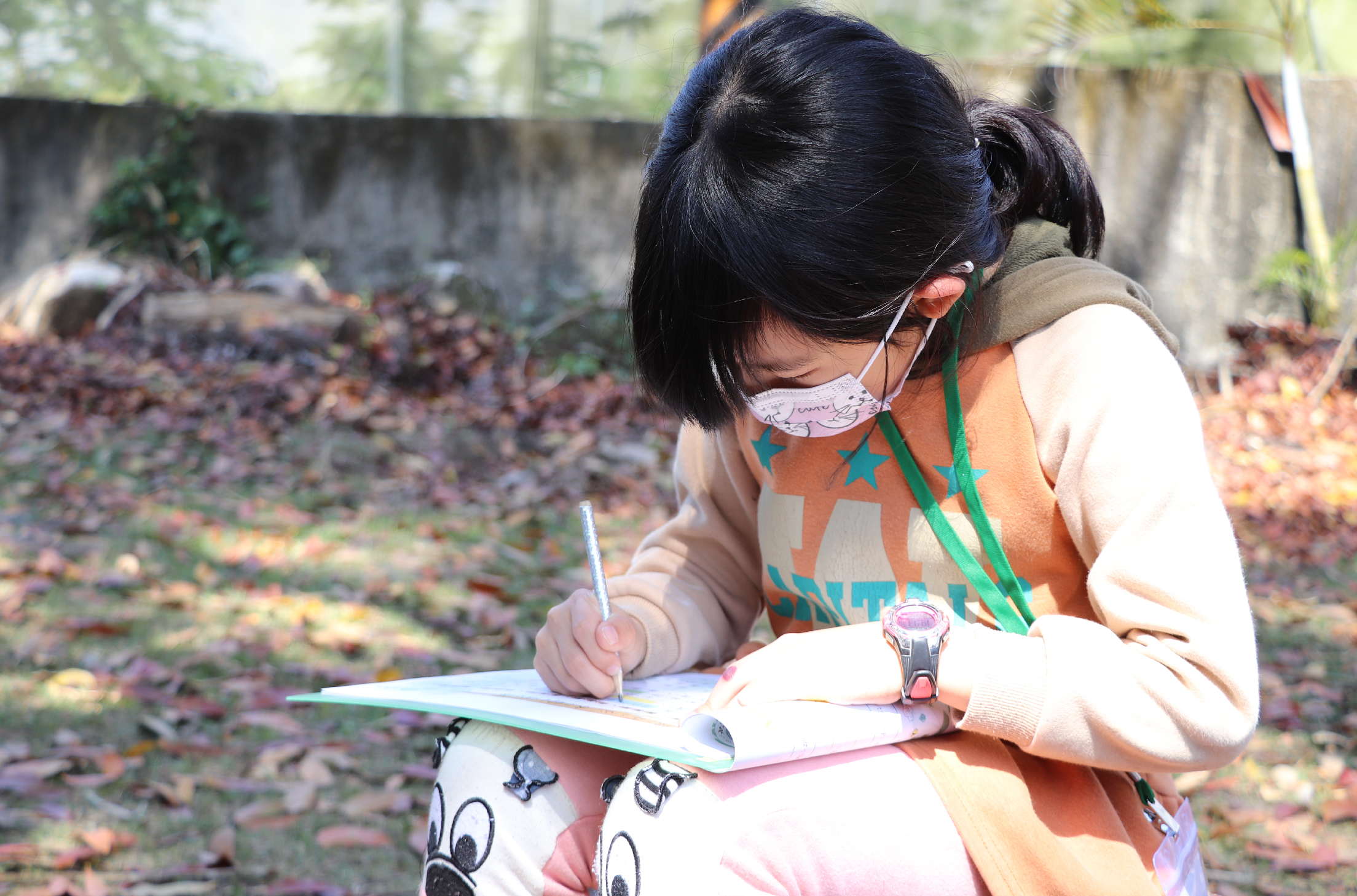 一名戴著口罩的女孩坐在戶外，穿著橙色上衣，正在低頭認真地在筆記本上寫字。背景是落葉遍地的庭院和灰色牆面，她手持藍色筆，專注於書寫作業或繪畫。