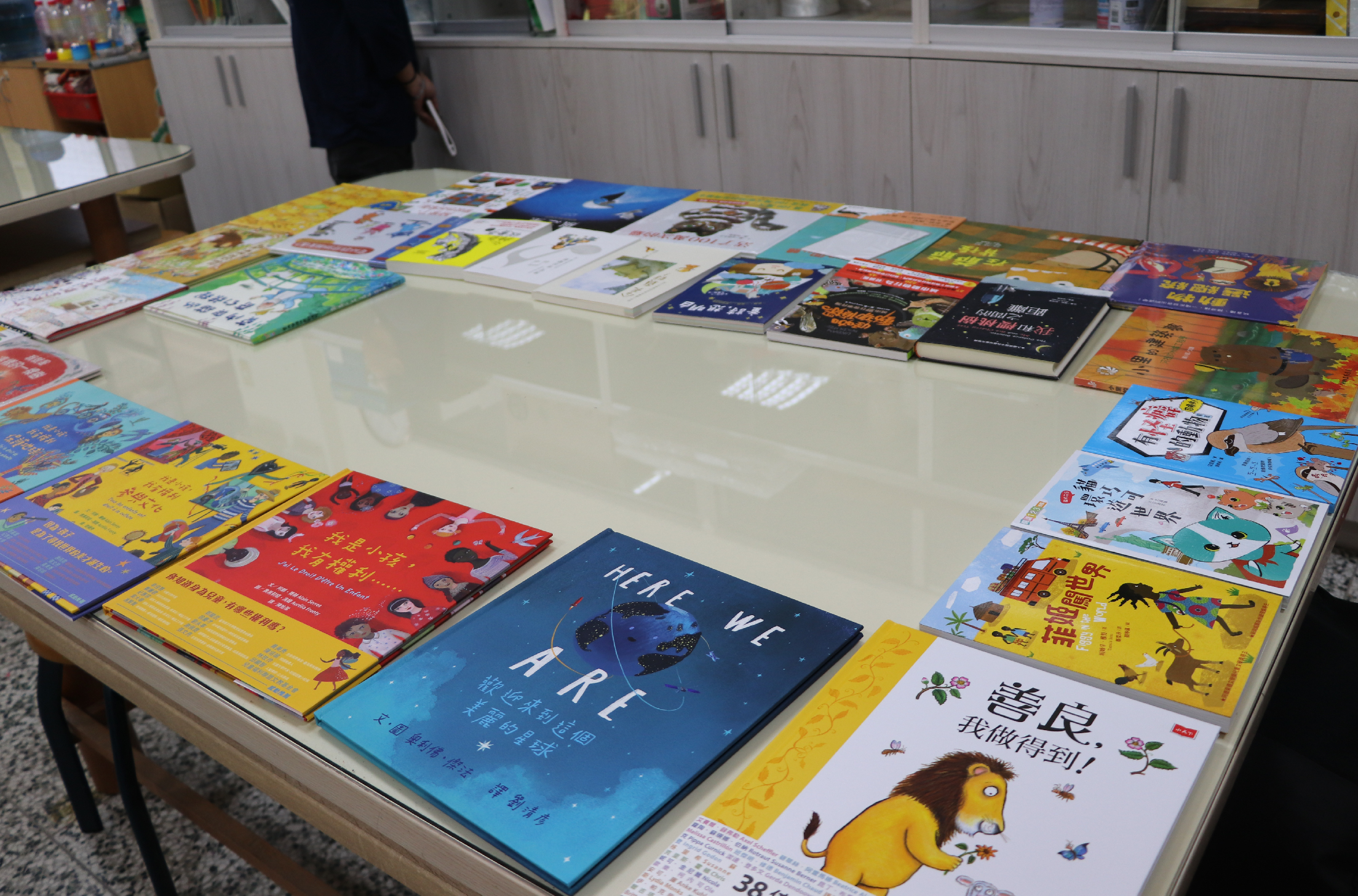 圖書館展示桌上排列著多本中文童書和繪本，色彩繽紛，包含黃色、紅色、藍色等多種書籍。背景為白色櫃子，桌上展示了約三十本不同主題和風格的兒童書籍，供閱讀和選購。
