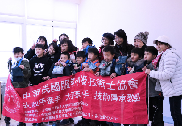 一群大人和小孩在室內合影，共同舉著寫有「中華民國服裝設計師協會」和「衣啟年華 技術傳承教學」等文字的紅色布幡，展示傳統服飾文化推廣活動的成果。