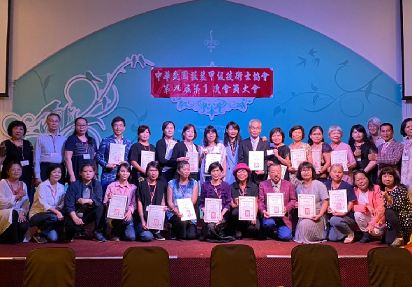 一場頒獎典禮，舞台背景為藍色漸層設計，紅色橫幅標語居中。約四十位獲獎者穿著正式服裝，分兩排站立舞台上，手持證書合影。現場布置典雅，反映正式的表彰活動。