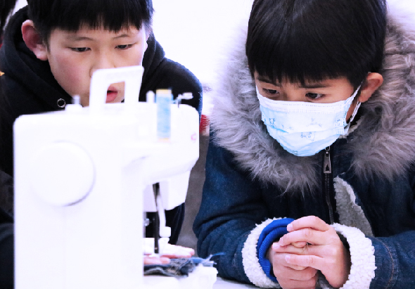兩名戴著口罩和手套的兒童在室外操作科學儀器，左側男孩專注於白色機器，右側女孩低頭觀察手中的實驗用具，展示兒童參與科學教育活動的情景。