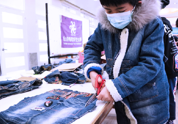 一位戴著口罩和黑帽的女性，穿著牛仔夾克在室內工作台前，使用紅色工具對牛仔褲進行改造或修飾。桌上擺放著多件牛仔服飾和配件，背景可見電腦螢幕和紫色海報，呈現手工藝或服裝改造的工作場景。