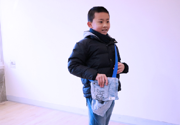 一名年幼男童穿著深藍色羽絨外套，背著藍色背包，手持印有文字的淺藍色牛仔布袋子，轉身面向鏡頭微笑。背景為白色牆面。