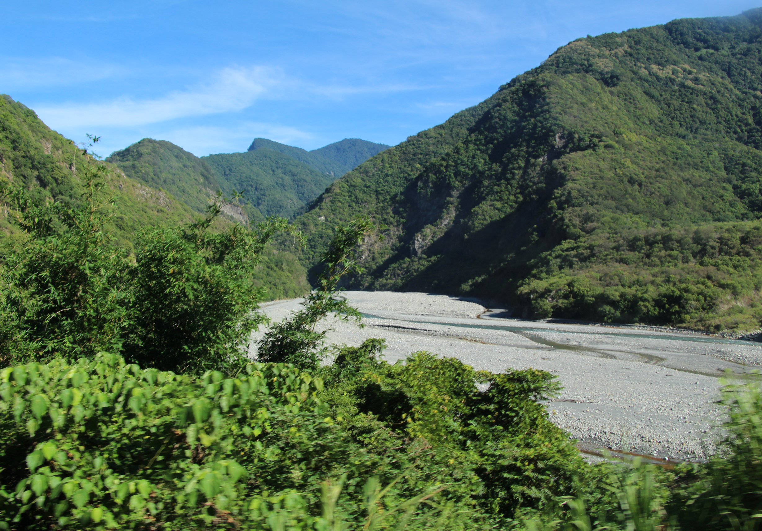 壯麗的山谷風景，兩側陡峭的綠色山峰覆蓋密集森林，中間流淌著灰白色的河流。清澈藍天映襯著群山，前景為青綠的灌木叢。