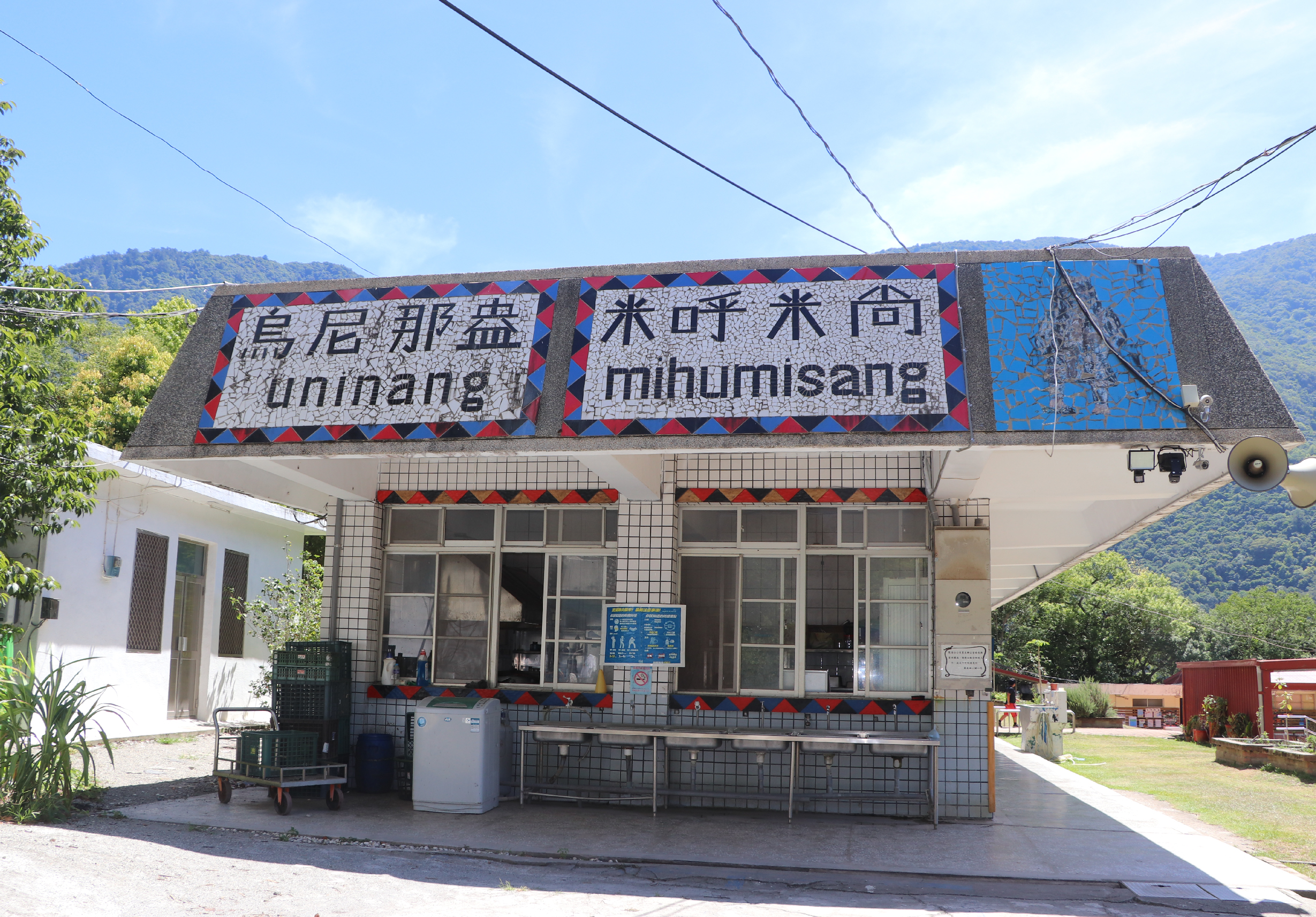 烏尼那盞（uninang）米竹米商店鋪，白瓷磚外牆搭配紅藍幾何裝飾邊框，招牌以中文及羅馬字標示店名，玻璃門窗前放置冰箱及推車，背景為青翠山景，典型台灣原住民社區商店。