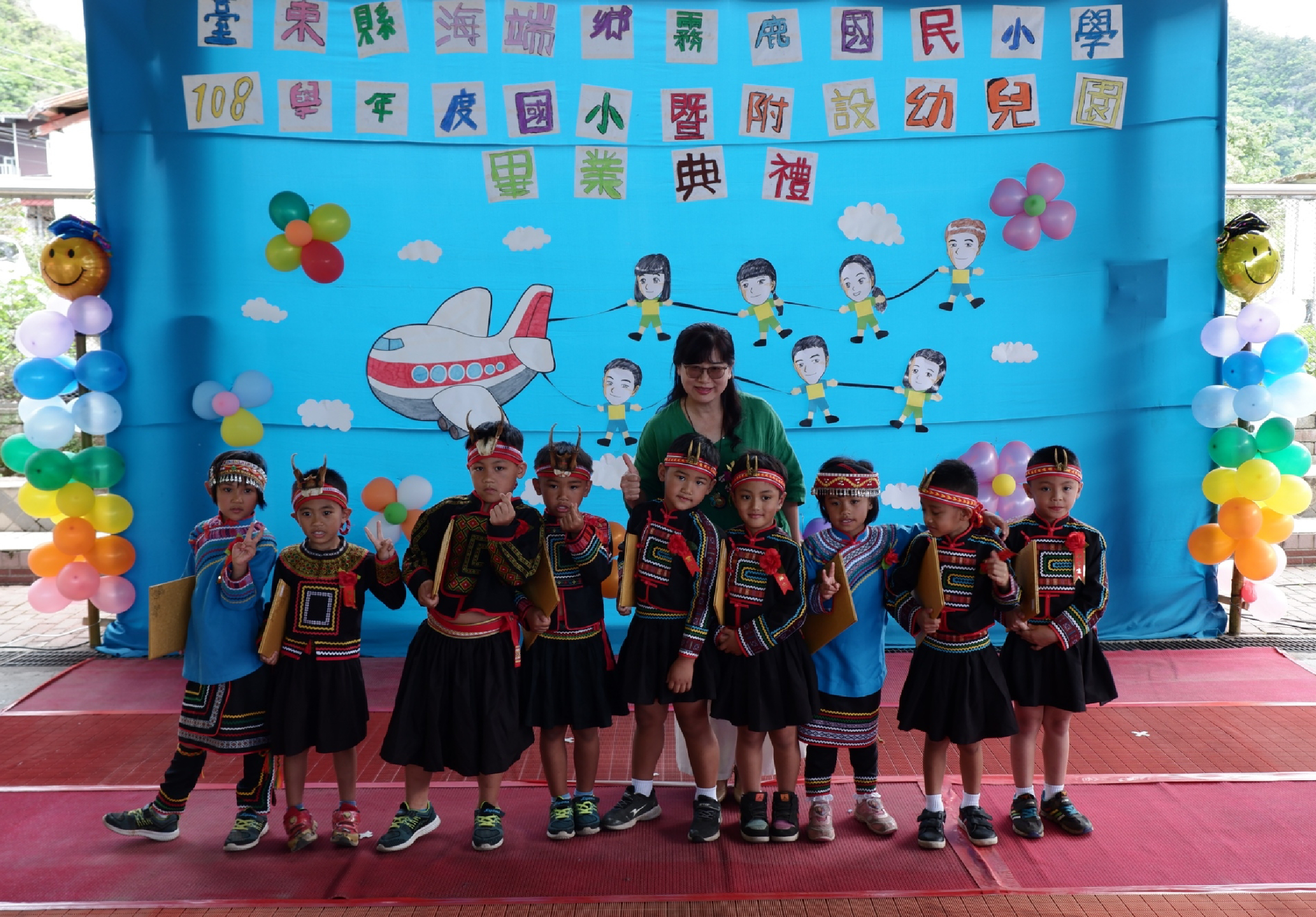 一位女老師與九位穿著傳統原住民服飾的小學生站在布置精美的舞台上。背景是藍色布景，上方有中文標題和插畫，兩側裝飾彩色氣球。舞台地面為紅色，學生們擺出歡樂姿勢合影。