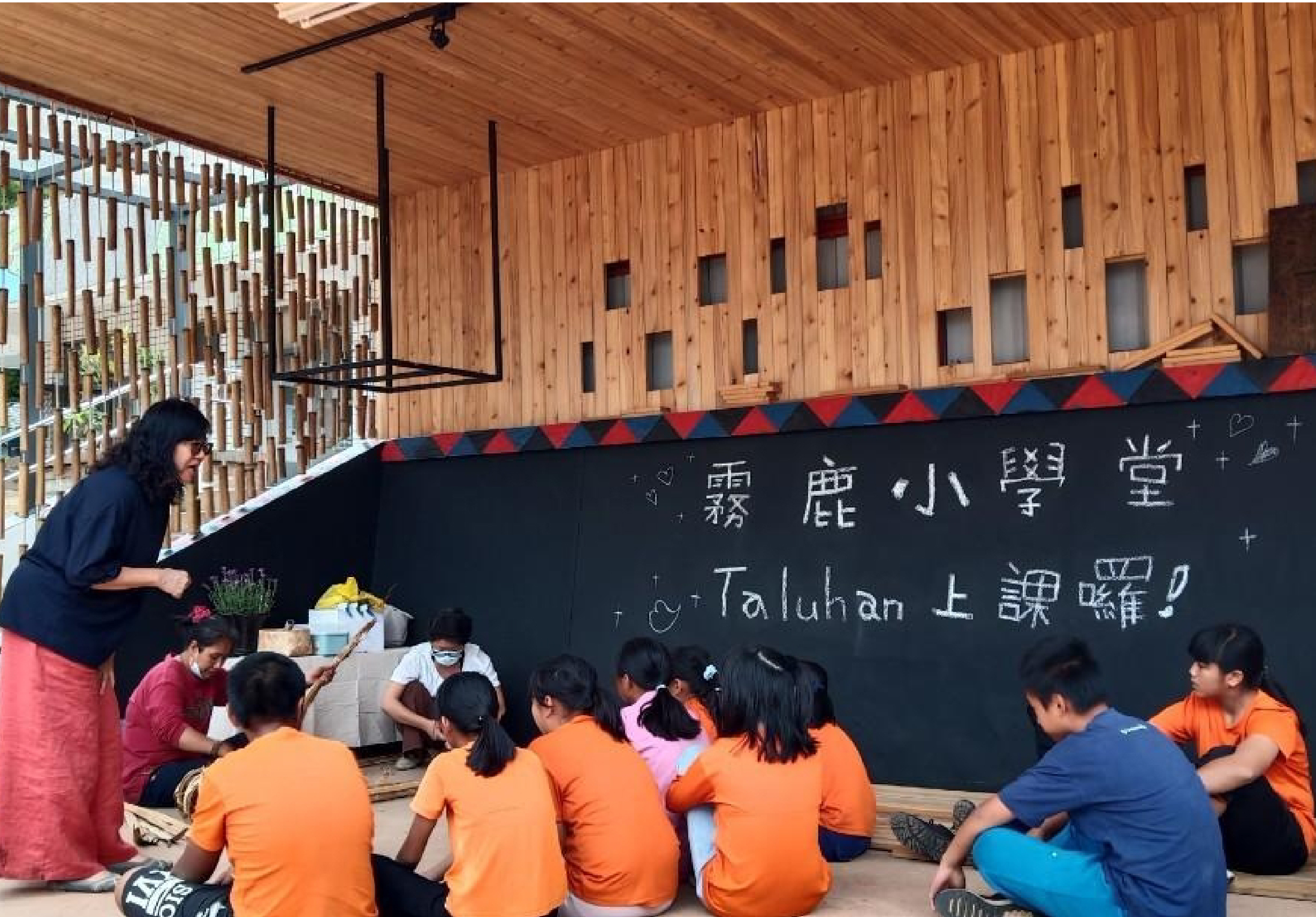 霧鹿小學堂教室內，穿著橙色制服的學童圍坐在黑板前，聆聽穿著深藍色服裝的教師講課。黑板上以中文和英文書寫著課程內容。木質建築搭配紅藍三角旗裝飾，營造溫暖的學習環境。
