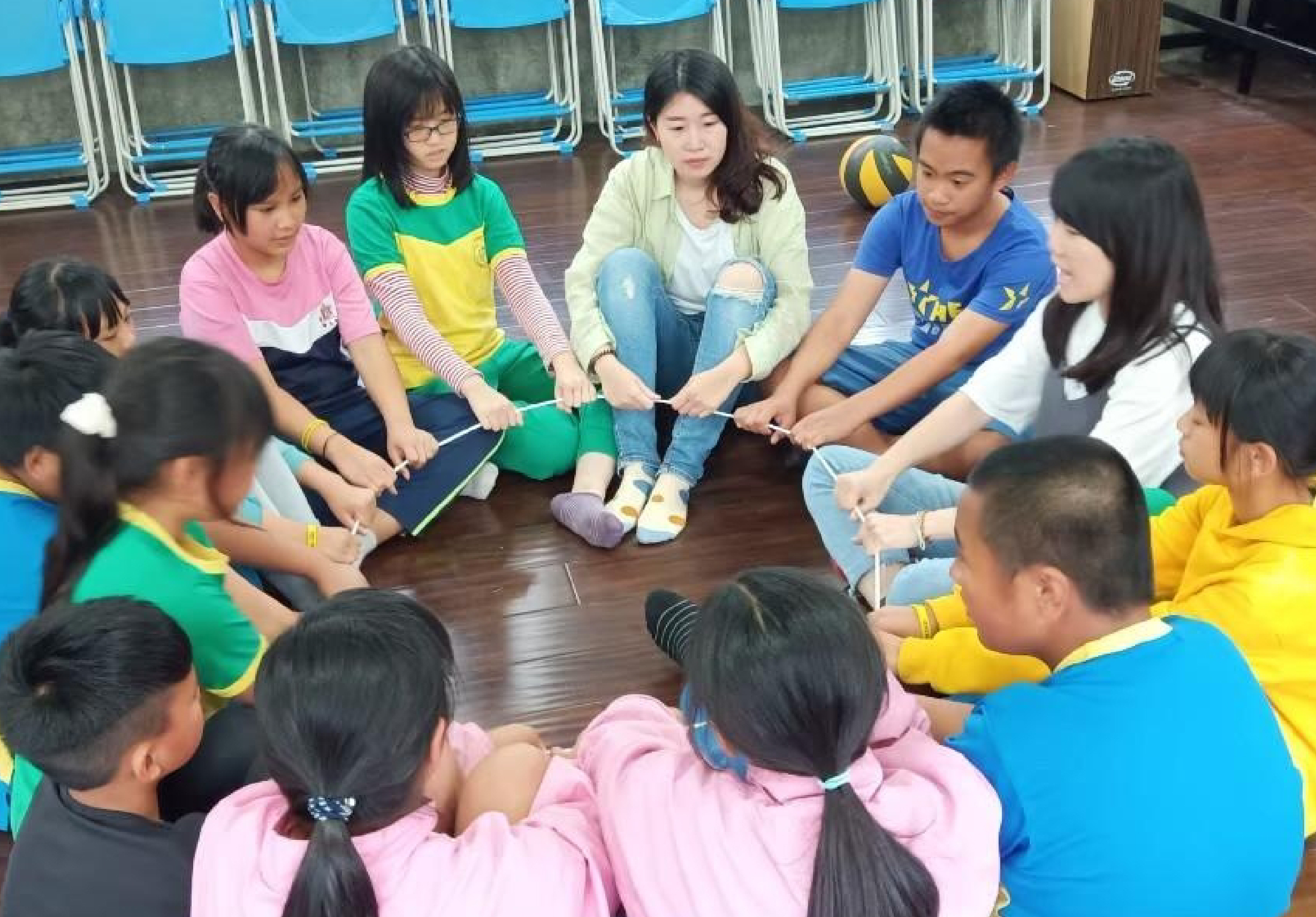 一群學生圍坐在木質地板上，手持繩子進行團隊建構活動。教室內有藍色折疊椅和排球，學生穿著不同顏色的運動服，專注參與互動遊戲，展現團隊合作精神。