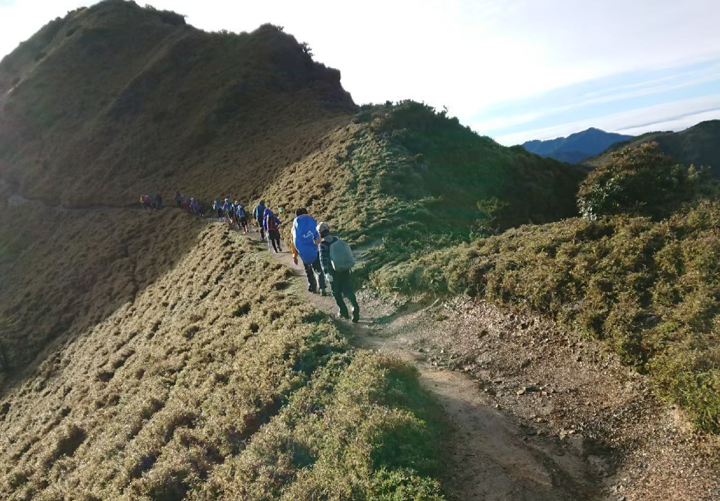 一群登山者沿著山脊步道行進，穿著藍色和深色衣著，背著登山包。步道兩側是裸露的土壤和稀疏植被，遠方可見山脈和藍天。場景呈現登山隊伍在高海拔山區進行集體登山活動。