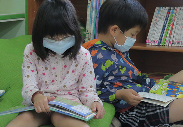 兩名戴著口罩的小孩坐在綠色地墊上，在圖書館內閱讀彩色圖書。背後是書架，放滿各類書籍。兩人專注地翻閱手中的繪本，進行閱讀活動。