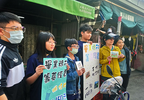一群佩戴口罩的學生在街道活動中舉著宣傳牌，展示環保或社區服務的標語。多名學生穿著不同顏色的衣服，在戶外參與集體倡議活動，背景可見綠色帳篷和店鋪。