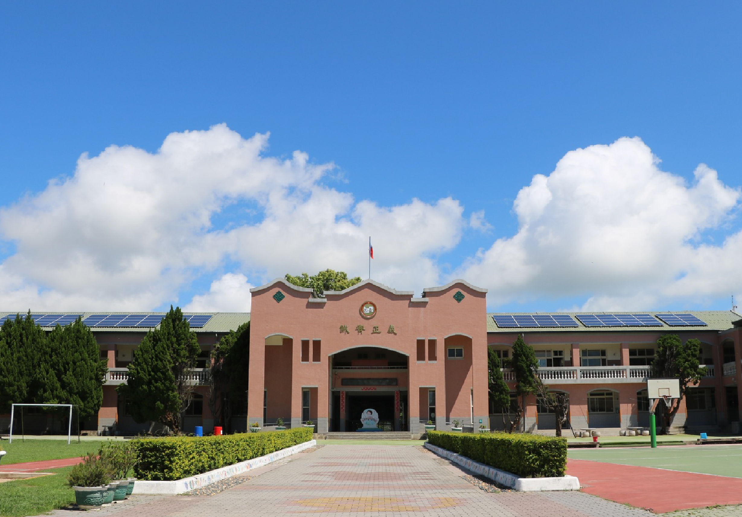 一座紅磚校舍建築物，正面有大型拱形入口和旗幟，兩側為對稱的翼樓。校園前方有寬闊的紅磚步道和綠化草坪，左側可見足球門框。屋頂裝設太陽能板。晴朗天空飄浮白雲，景象寧靜祥和。