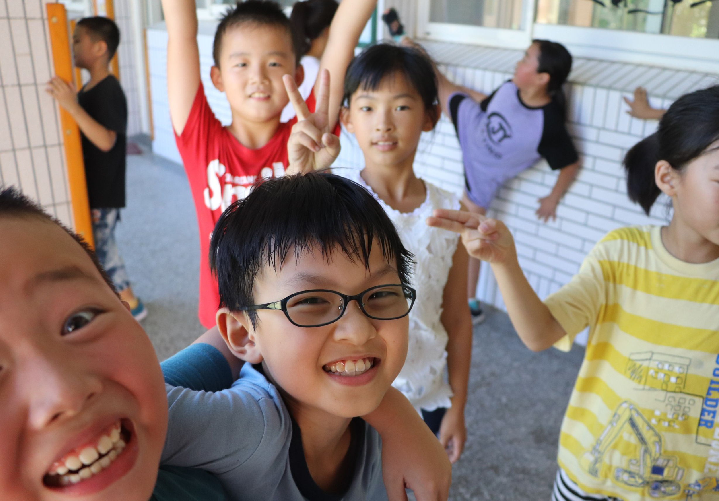 一群小學生在走廊裡開心地聚集，穿著紅、黃、紫、藍等彩色衣服。前景一名戴眼鏡的男童燦爛微笑，身旁女童做出勝利手勢，背景還有多名學生揮手互動，氣氛歡樂溫暖。