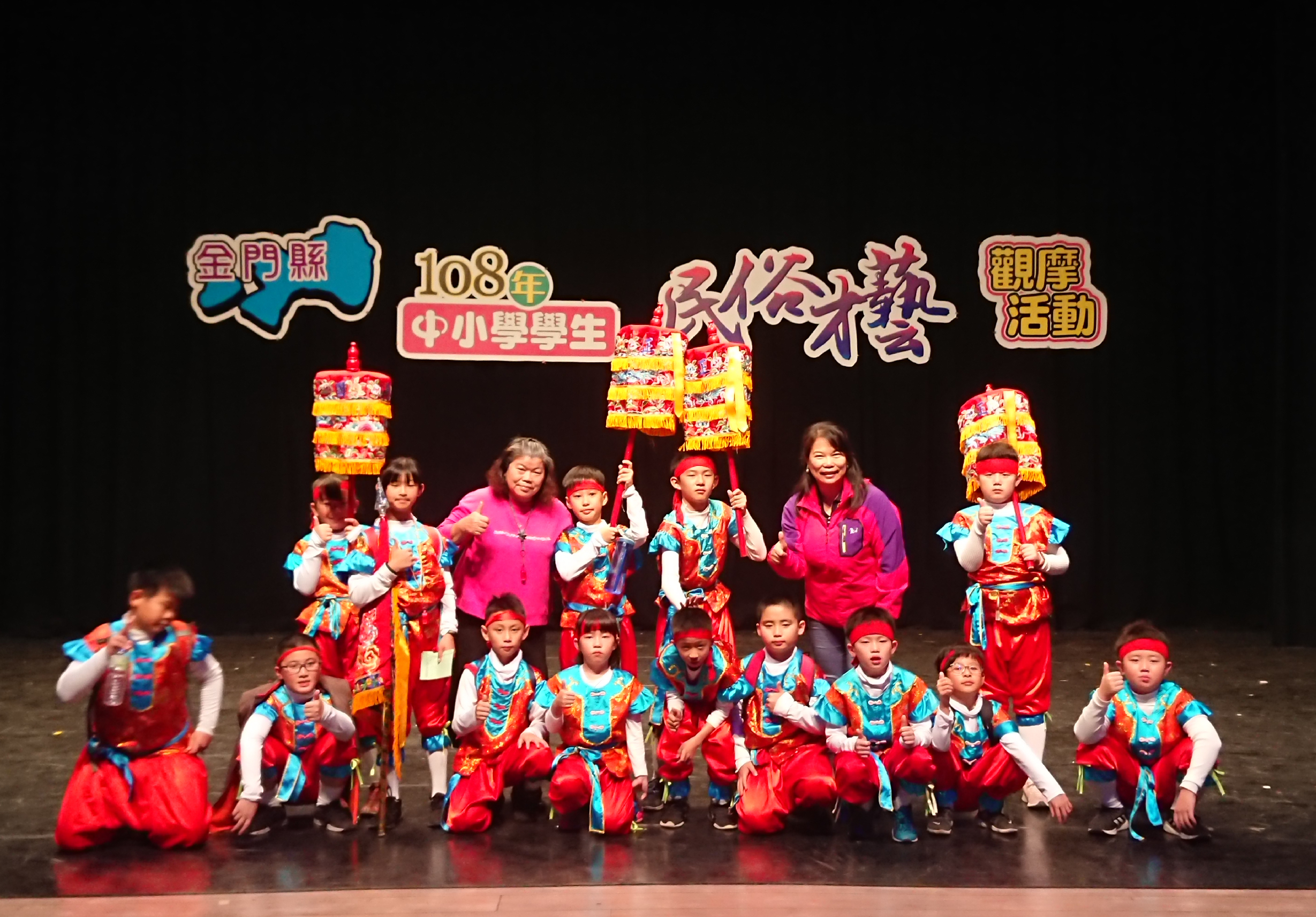 金門縣108年中小學學生「慶祝農曆新年」舞蹈活動現場。舞台上約20位穿著紅藍相間傳統服裝的學生，配戴紅色頭飾，兩位教師指導，後方懸掛彩色花燈，展示傳統新年慶祝表演。