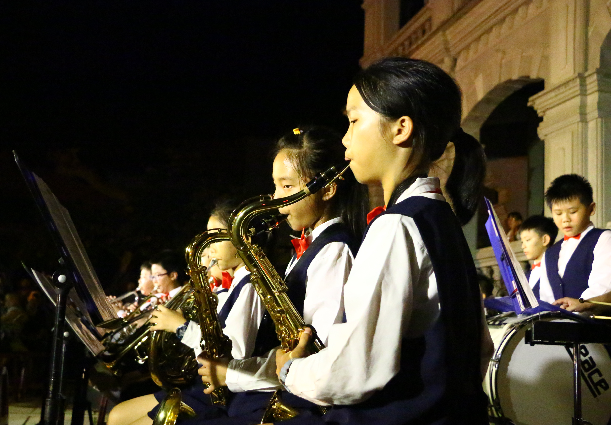 夜間戶外演出現場，身穿海軍藍白相間制服、配戴紅色領結的學生樂手們站成一排，專注地演奏薩克斯風。樂譜架前的演奏者們集中精神表演，身後古典建築在燈光映照下呈現溫暖金色，營造莊重的音樂會氛圍。