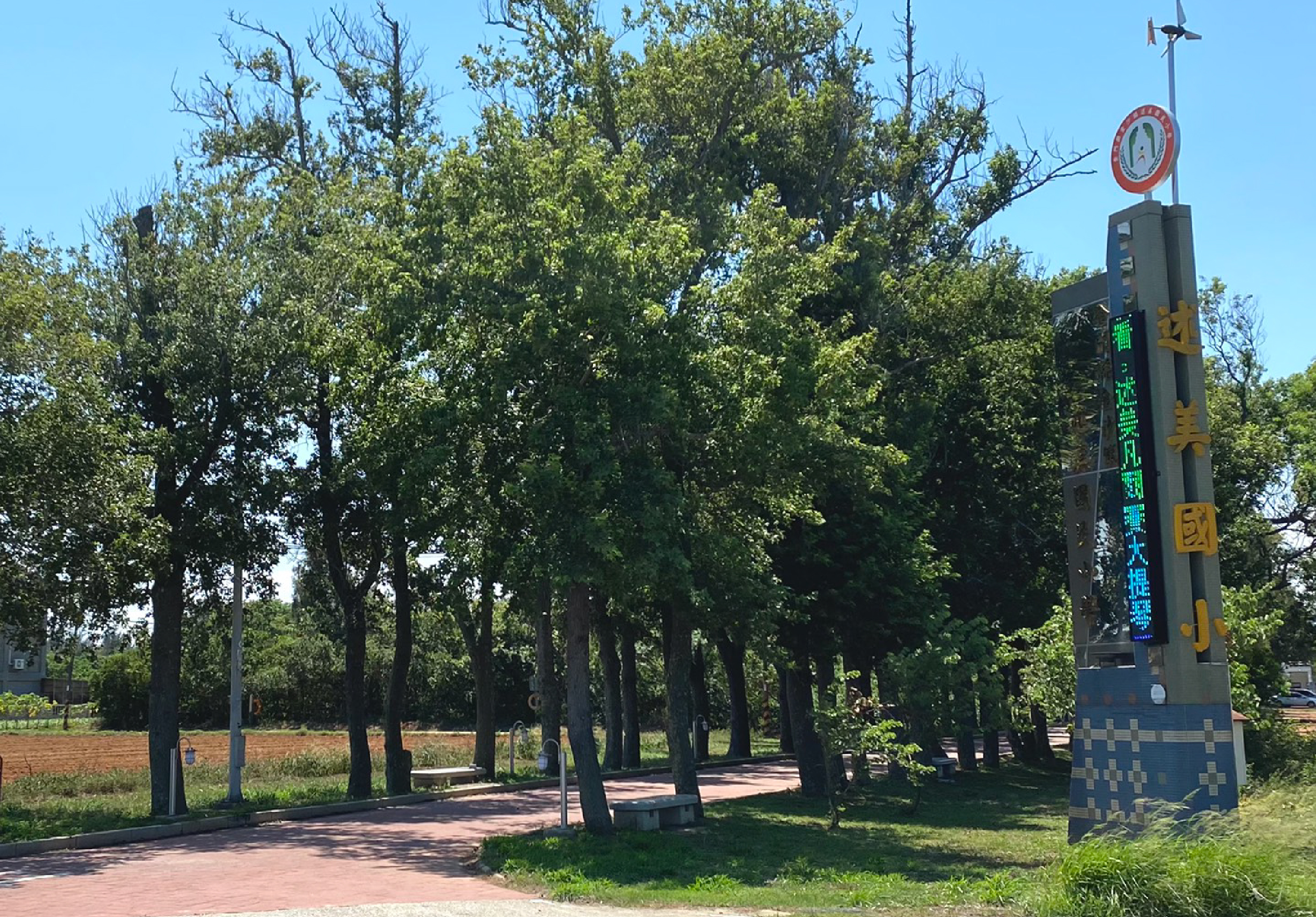 公園內林蔭步道兩旁種植高大樹木，右側豎立一座藍色基座的時間溫度顯示塔，塔頂有紅色圓形標誌。地面為磚紅色步道，遠方可見田地和建築物。