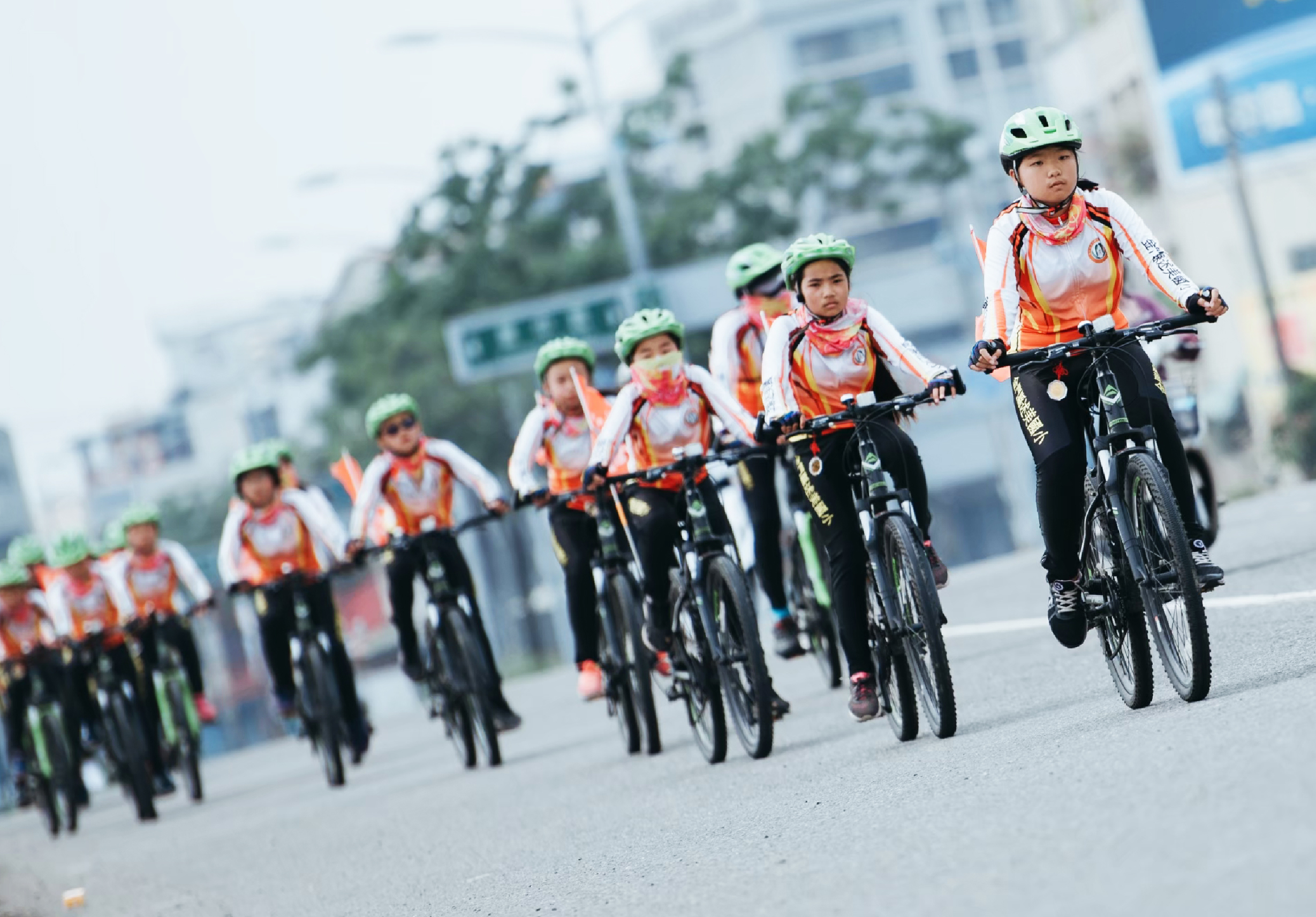 一隊穿著白色和橙色運動服的年輕騎手在城市街道上騎自行車遊行。他們都戴著綠色安全帽，騎著黑色自行車，背景是高樓大廈。這是一場大型自行車集體活動或宣傳活動。