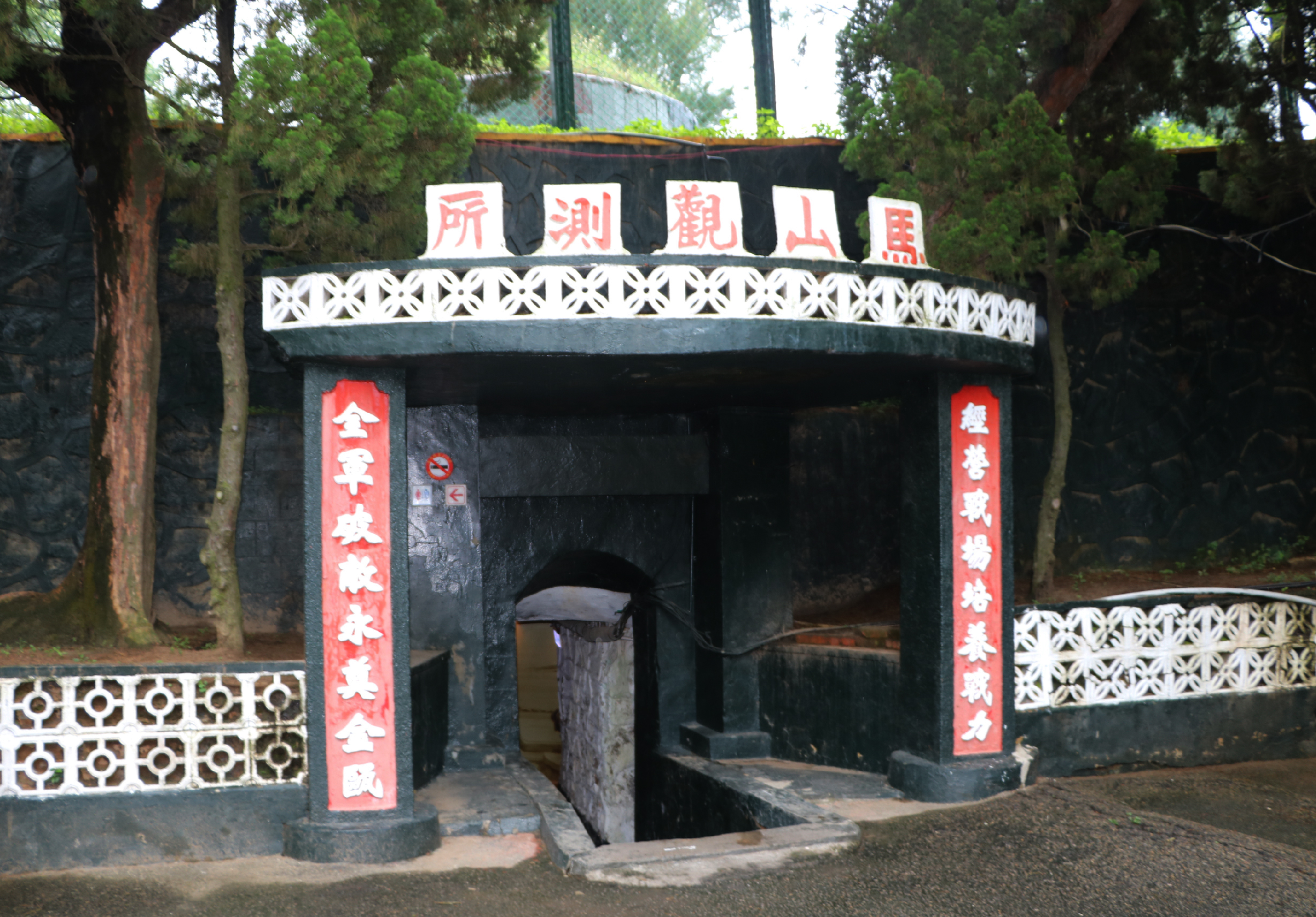 古典中式建築牌樓，黑色石材構造，頂部白色裝飾邊框，上方五塊白底紅字匾額，兩側紅色對聯，下方拱形石門入口，周圍環繞老樹，具有濃厚東方傳統風格。