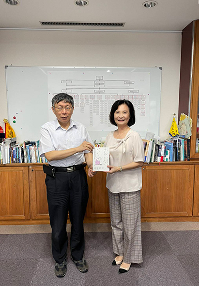 辦公室內，一名男性和一名女性並肩站立，女性手持一本書籍。身後白板上繪有組織結構圖，左右兩側書架上放置多本書籍和裝飾品。