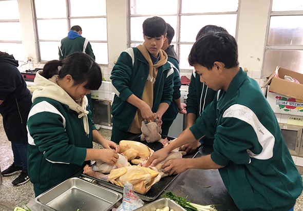 四名穿著綠白相間校服的學生在教室內進行烹飪實習，圍繞著料理台合作處理食材，講師在中央指導。明亮的教室窗戶和後方的儲物架清晰可見。