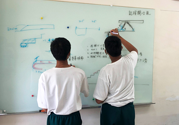 兩名穿著白色短袖上衣的學生在whiteboard上討論物理槓桿原理的問題。左邊學生觀看，右邊學生用手指著board上的槓桿示意圖進行講解。board上有藍色的槓桿裝置圖、計算過程和中文標註。