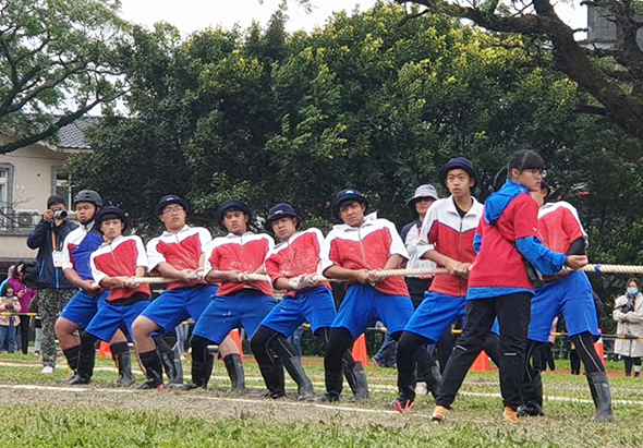 一群穿著紅藍運動服的小學生在草地上進行拔河比賽，約十五人排成一列用力拉著粗繩。背景可見樹木、房屋和觀眾，展現校園運動會的熱鬧氣氛。