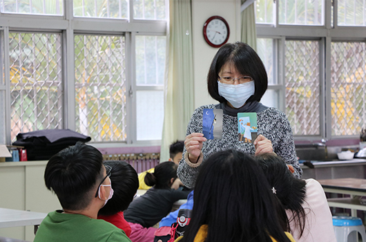 教室內，戴著口罩的女性教師手持教材，站在圍坐成圈的學生面前進行教學。窗戶後方可見校園景致，教室內有時鐘、暖氣管和教學用具。