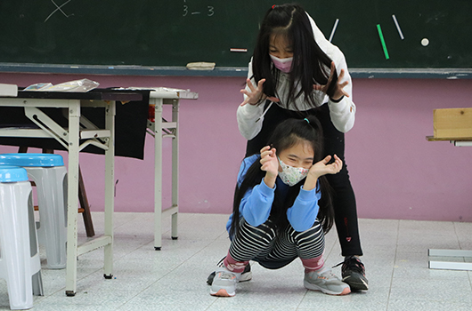 教室內，一位女學生站在後方，雙手搭在前方蹲坐男學生的肩膀上。兩人均戴著口罩，背景是粉紅色牆面和綠色黑板。地板上有教學用具。