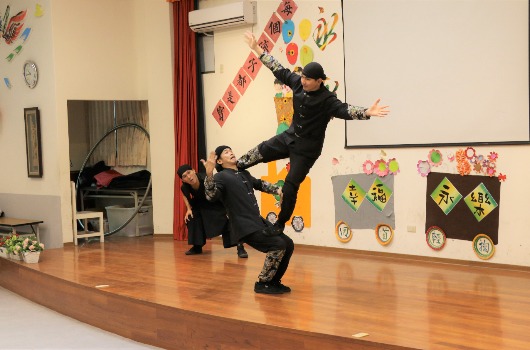 室內舞台上，一位穿著黑衣的表演者做出武術動作，一隻腳高高抬起。舞台背景裝飾著紅色布簾、彩色紙製裝飾和中文字牌。舞台下坐著觀眾觀看表演。