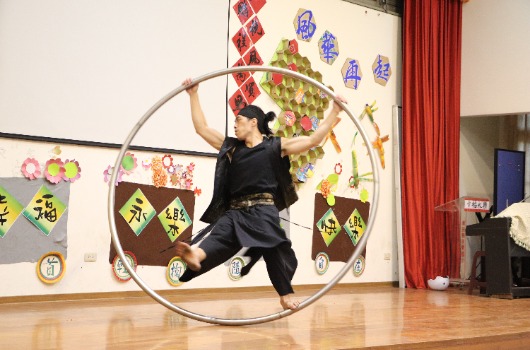 身穿黑色服裝的表演者在室內體育館舞台上，跳躍著轉動一個大圓形呼啦圈。背景裝飾著紅色布幔和傳統亞洲風格的彩色掛飾，地板為木質。表演展示了體操或雜技技能。
