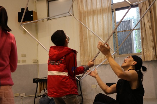 室內教室活動中，穿著紅色背心的兒童舉起竹竿，成人女性協助指導。窗戶透進自然光，牆上有電視螢幕。這是一堂互動式的兒童學習或運動課程。