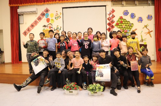 一群小學生在教室內合照，背景布置有紅色布幕、聖誕裝飾和春節佈置。學生們穿著冬季服裝，有人拿著獎狀和作品，站在木製舞台上，前方放有盆栽。這是一場校內慶祝或頒獎活動的集體合照。