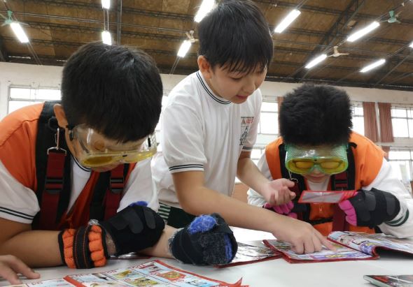 三位穿著橙色安全背心和防護眼鏡的小學生，在室內場館的工作台前進行手工藝活動。中間男童正指導左右兩側的學生一起製作或觀察手工作品。桌上散放著彩色紙張和製作材料。